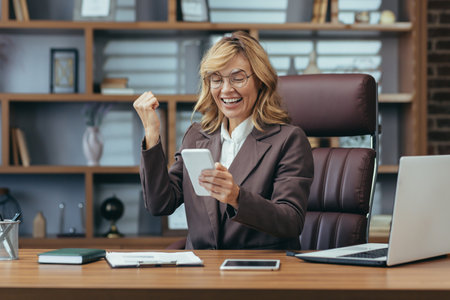 Cheerful mature businesswoman with glasses celebrates a successful deal or news while working remotely on her smartphone in a home office.の写真素材