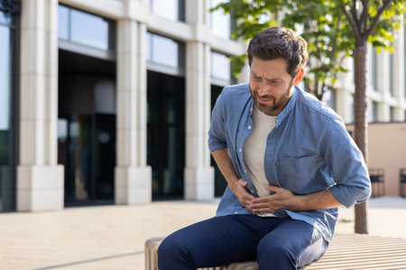 A mature bearded businessman in casual attire appears distressed with a stomachache outside an...