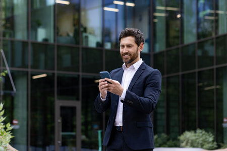 A professional man in a tailored suit enjoys a break outside, browsing on his phone with a content smile.の写真素材