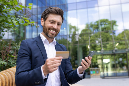 Confident male executive managing finances with phone and card, standing by an office building...
