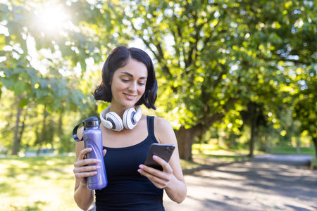 Fitness-focused female enjoys a break in the park, checking her phone with water bottle and...