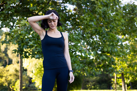 Young woman jogging and doing sports in the park, standing in sportswear and holding her head with...
