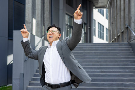 Joyful businessman celebrating success outside office building, pointing upwards with excitement and achievement in urban setting.の写真素材