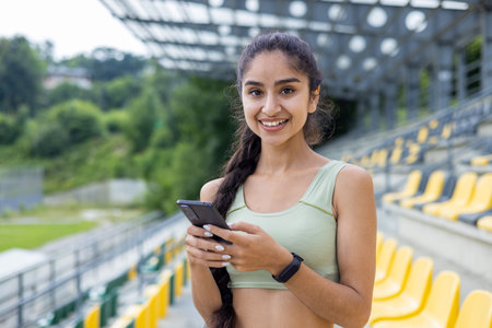 Smiling young Indian woman in a tracksuit adjusts her smartwatch during a sport and fitness routine...