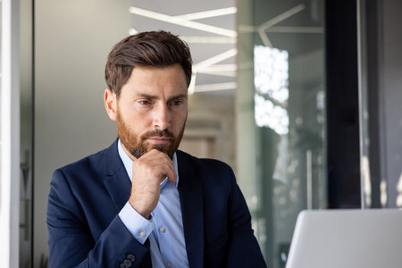 Close-up photo of a young serious male businessman sitting focused in the office at a laptop and thinking about information, leaning his head on his hand.の写真素材