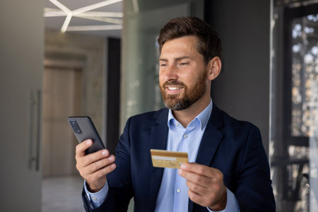 Close-up photo of a young businessman standing inside an office center in a suit, holding a credit...