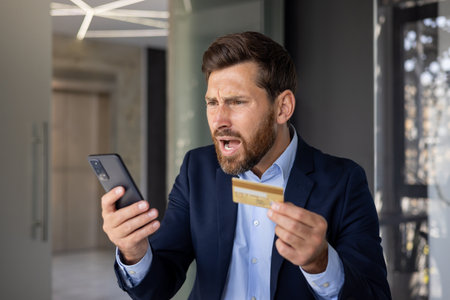 Close-up photo of angry young businessman in suit standing in office, holding golden credit card in hand and looking disappointedly at mobile phone screen.の写真素材
