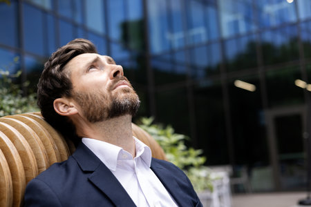 Close-up photo of a young pensive and tired businessman sitting outside an office building, leaning on a bench, looking up, upset and disappointed.の写真素材