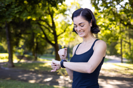 Happy young woman is doing sports and running in the park, standing looking at the smart watch and...