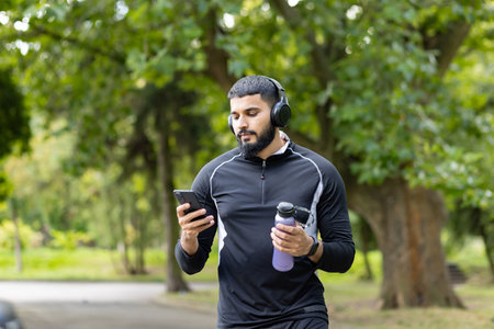 A fit man in sportswear takes a break in a lush park to use his smartphone and drink water,...