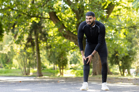 A man in sportswear holds his knee in pain while exercising in a park setting, showcasing the...