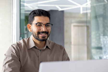 Smiling Indian businessman in an office setting actively engaged on his laptop, exuding...
