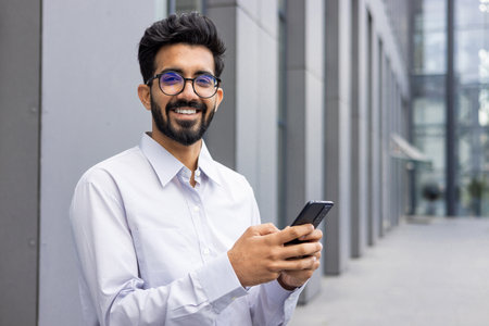Portrait of a smiling and happy young Indian man in a white shirt and shorts standing near an office center, holding a phone and smiling at the camera.の写真素材