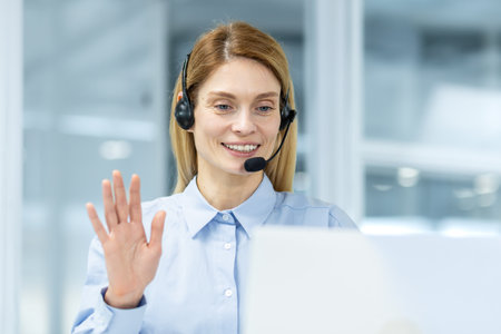 Friendly businesswoman with a headset making a cheerful greeting gesture during a video conference in an office.の写真素材