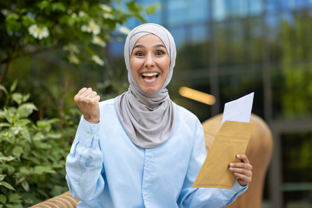 Vibrant image of a joyful Muslim woman wearing a hijab, exuberantly celebrating while holding an envelope outdoors with a blurred green background.の写真素材