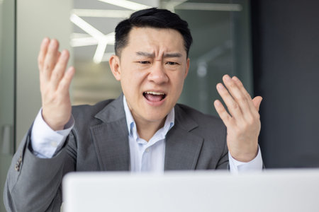 Annoyed Asian man in a suit making gestures on a stressful video chat at work with a laptop in front...