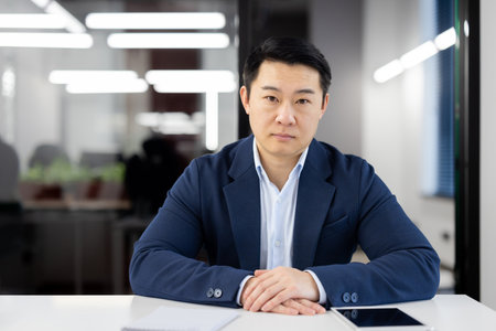 Portrait of a confident Asian businessman seated at a table in a well-lit, contemporary office environment, looking at the camera with focus.の写真素材