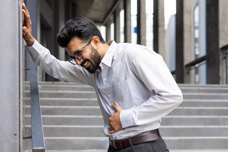 Young Indian man suffering from stomach pain, holding hands to abdomen, standing outside office building, having panic attack and stress about work.の写真素材