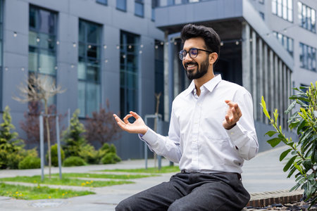 Smiling and relaxed young Indian male businessman sitting on bench in lotus position near offices, meditating and resting with eyes closed.の写真素材
