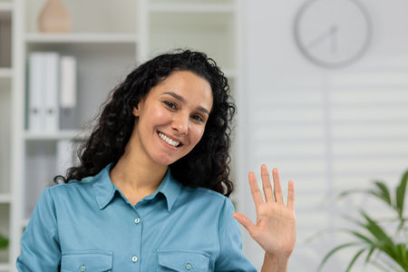 Smiling woman in a casual shirt waving hello during a video call, providing an engaging webcam view POV in a home setting.の写真素材