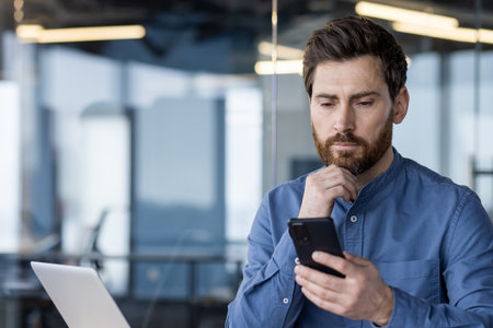 Professional businessman in a modern office setting, intently using a smartphone while working on a laptop.の写真素材