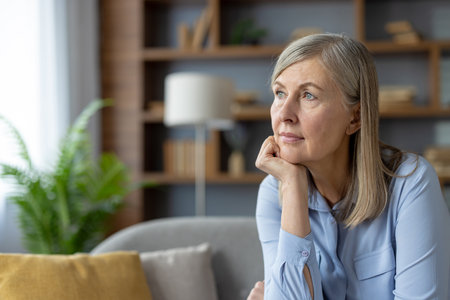 A serene mature woman in a blue shirt stares thoughtfully while sitting in a well-lit living room. The peaceful atmosphere highlights her pensive mood.の写真素材