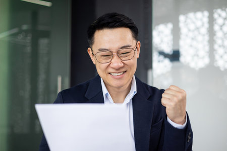 Asian mature man smiling in office, holding papers and celebrating success with raised fist, signifying achievement and happiness.の写真素材