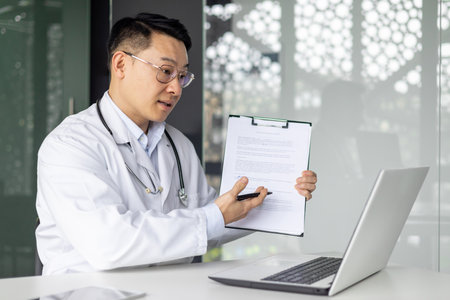 Asian mature male doctor explaining a medical report during a video consultation using a laptop in a modern office.の写真素材