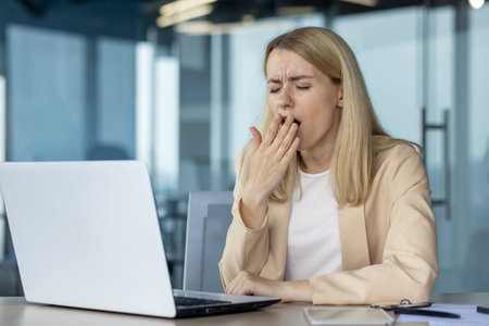 A blonde woman in a light suit feeling exhausted while yawning in front of her laptop in a modern office setting.の写真素材