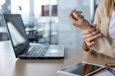 Businesswoman experiencing wrist pain while working on laptop in a modern office setting. Highlighting office ergonomics and workplace health.の写真素材
