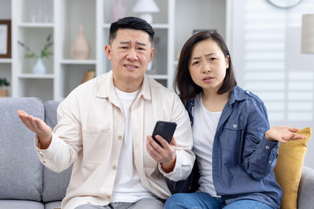 Confused couple sitting on a couch at home holding a smartphone, expressing frustration and...