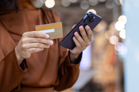 A person in a brown coat is shopping online, using a credit card and smartphone, in a shopping center with bokeh lights. They are making a payment and a purchase digitally with their mobile deviceの写真素材