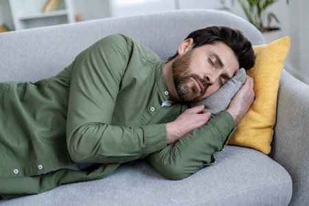 Bearded man peacefully napping on a gray sofa with yellow and gray pillows in a cozy living room environment.の写真素材
