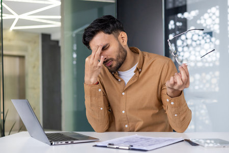 A businessman feeling stressed and frustrated while working at his desk in the office. He is rubbing his eyes and holding his glasses in one hand.の写真素材