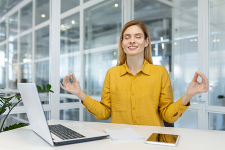 Relaxed businesswoman practicing meditation at her desk in a modern office to reduce stress and improve focus.の写真素材