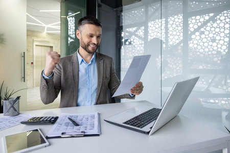 Successful businessman celebrating achievement in modern office, holding documents and using laptopの写真素材