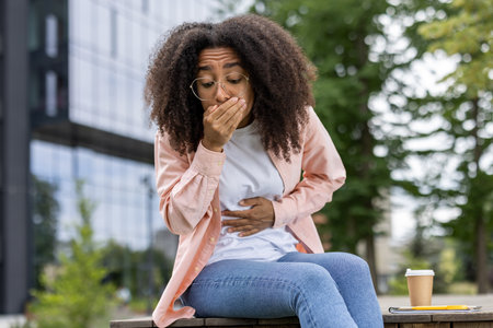 Young woman experiencing stomach pain and holding her hand over mouth. She is sitting outside in casual attire, indicating discomfort and possible nausea.の写真素材