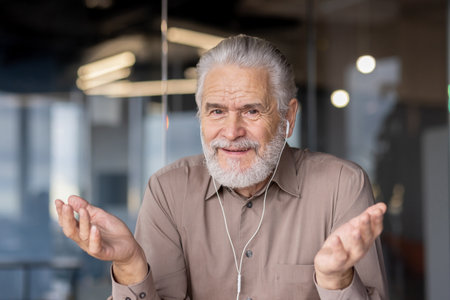Portrait of senior man with gray hair and beard wearing earphones gesturing while video chatting. Modern office setting with blurred background. Concept of communication and technology.の写真素材