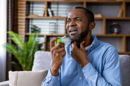 Man experiencing sore throat pain using throat spray for relief on couch at home. Concept of...