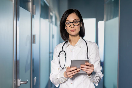 Confident female doctor with glasses and stethoscope holding digital tablet in modern hospital...