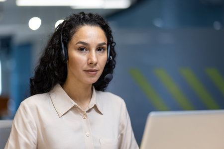 Focused professional woman with curly hair and headset working at desk in office environment. Modern...