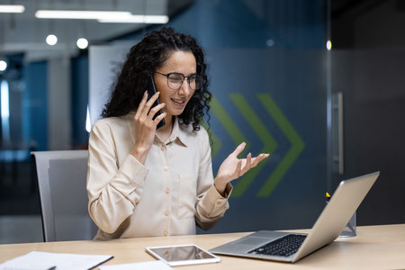 Confident businesswoman having phone call in modern office. Woman sitting at desk with laptop and...