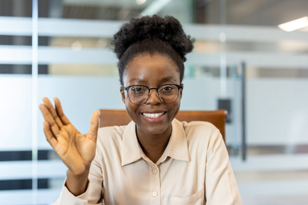 Smiling woman waving during video call in modern office. Friendly professional engaging in remote...