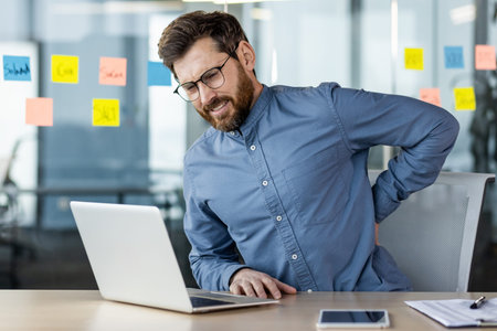 Businessman with glasses suffering from back pain while working with laptop at desk in modern...
