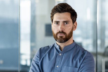 Close-up portrait of a young serious businessman in a blue shirt standing inside the office and...