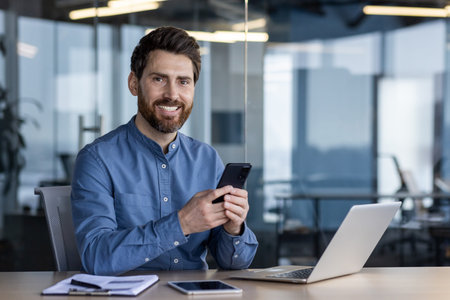 Portrait of a young successful male businessman sitting at a desk with a laptop in the office,...