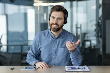 Portrait of a smiling young man in a headset and blue shirt sitting in the office at a desk and...