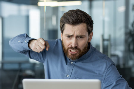 An angry young man in a blue shirt and with a beard is sitting in the office at a desk in front of a...