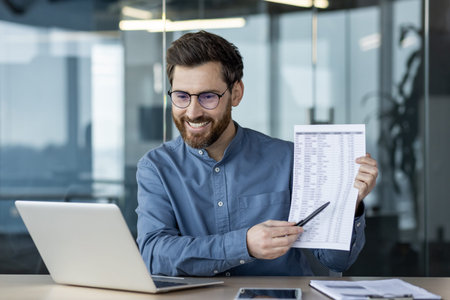 A smiling young man works in the office, sits at a desk in front of a laptop screen and shows data...