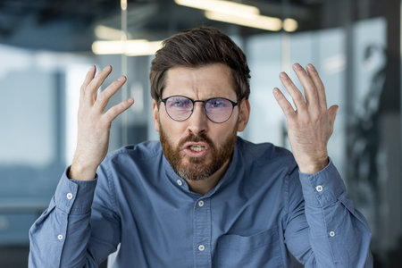 Close-up portrait of a young man wearing glasses sitting in a modern office and talking angrily and...